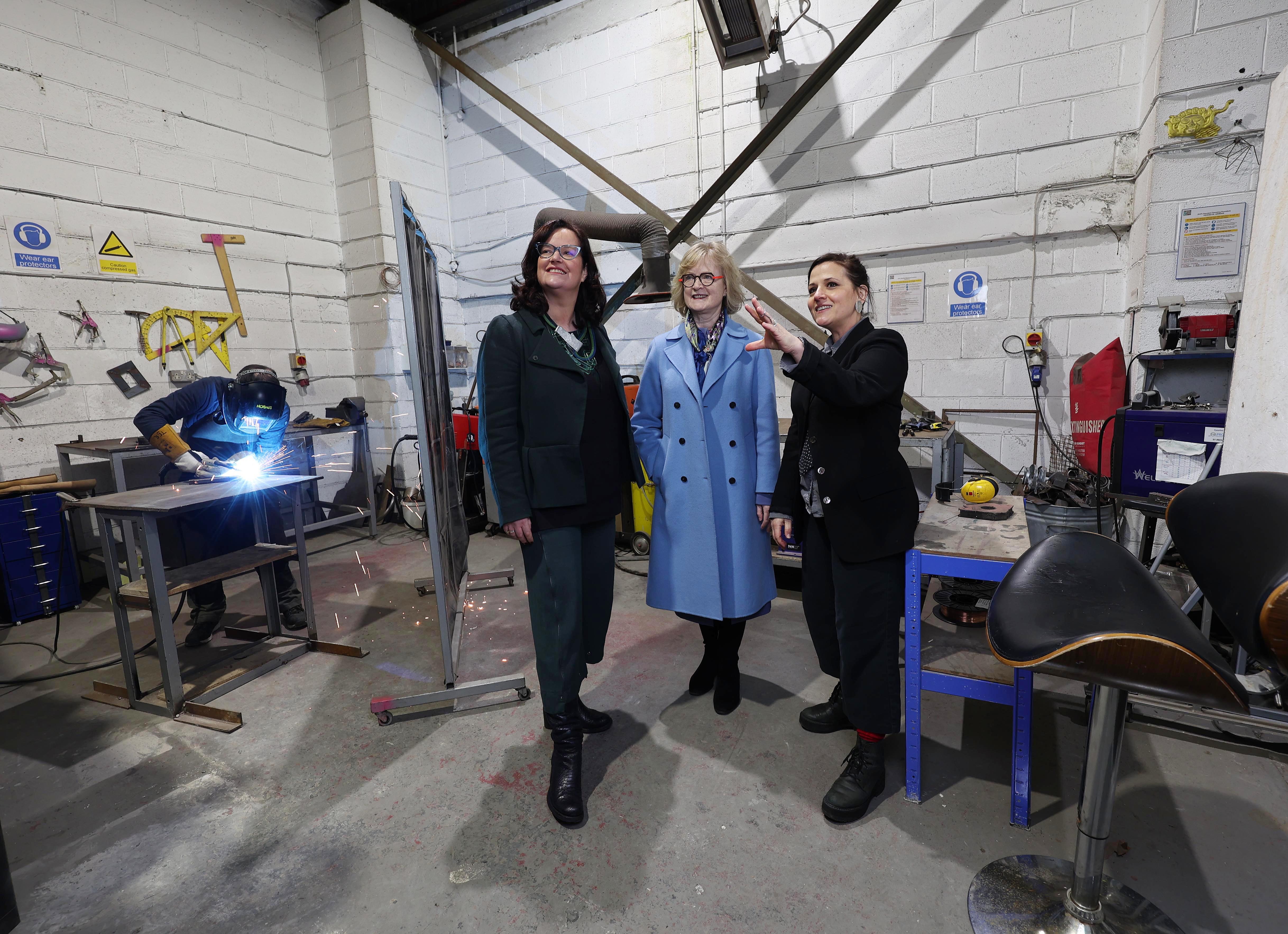 Three women standing in an industrial artists studio with a welder in the background