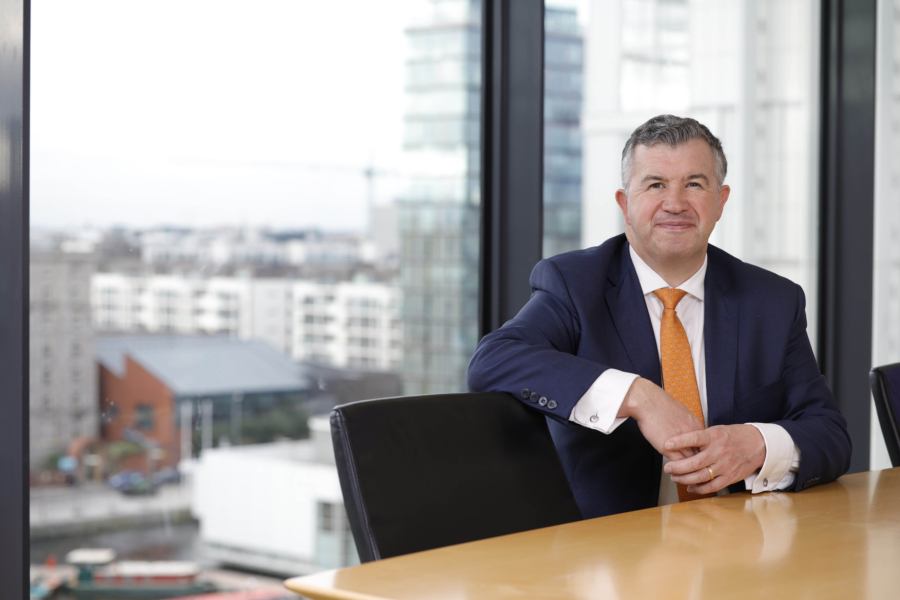 Smiling man in a dark blue suit, white shirt and orange tie, sitting at a desk in front of a window with a view of Dublin city