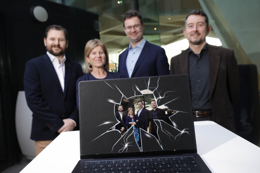 Four smiling people in business casual attire stand behind a laptop with a smashed screen, to symbolise 'disruption'