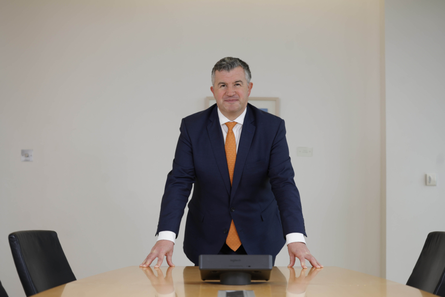 A businessman in a dark suit and orange tie leaning on a boardroom table.